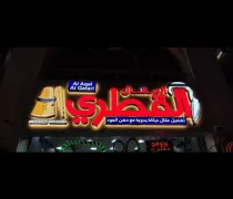 A brightly lit storefront sign featuring Arabic text. It showcases the name 'Al Qatari' with promotional phrases and a graphic logo