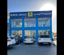 Blue and gray building with 'RUSIYA GROUP' sign, cars parked in front, bright sunlight in the background