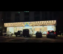 A brightly lit storefront displays 'RUSYA GROUP' in English and Arabic, surrounded by parked vehicles at night