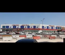 A commercial building with multiple shops in Arabic and English; traffic and construction barriers are visible in the foreground